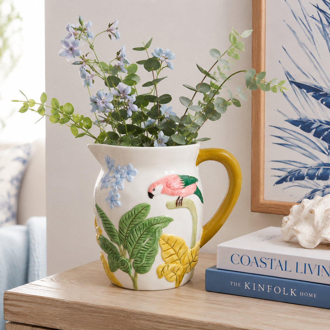A ceramic pitcher vase with a yellow handle and base, decorated with green tropical leaves, a pink flamingo, and blue flowers, filled with green eucalyptus and blue blossoms, styled on a wooden table next to books and a framed print.