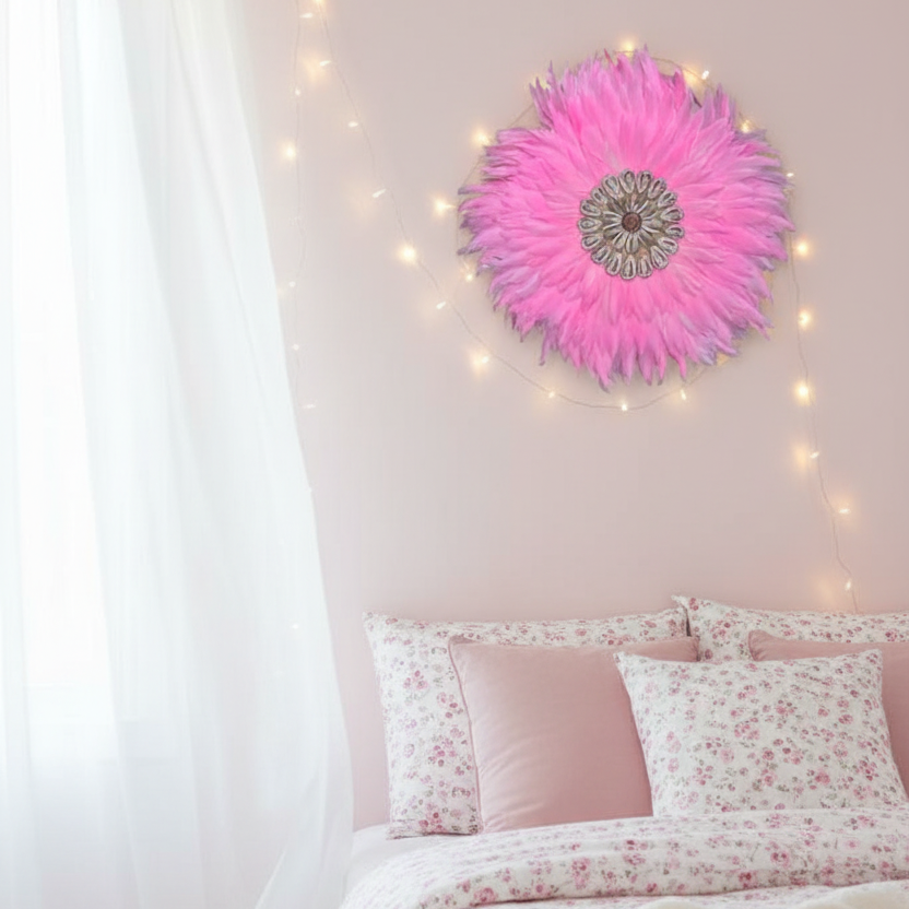 A styled bedroom corner with a bed dressed in pink and white floral bedding, a soft pink wall, string lights, and a large pink flower wall decor piece.