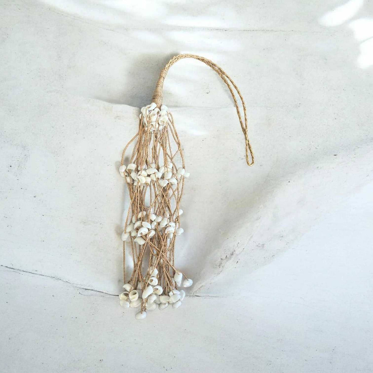 A bohemian-style tassel ornament featuring white shell beads strung on natural jute twine, hanging against a textured light gray concrete background.