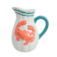 White pitcher with a pink crab design and blue handle 