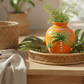 An orange ceramic vase with painted palm tree designs sits on a woven tray on a wooden table, styled with green plants and a beige cloth in a bright indoor setting.