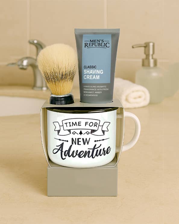 A boxed grooming gift set on a beige countertop in a bathroom setting, featuring a white ceramic mug with the phrase 'TIME FOR NEW Adventure,' a gray tube of Men's Republic shaving cream, and a shaving brush resting on top of the box.