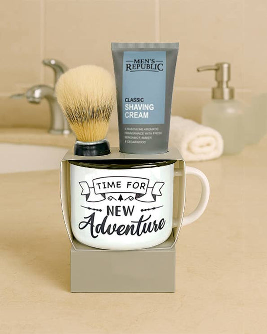 A boxed grooming gift set on a beige countertop in a bathroom setting, featuring a white ceramic mug with the phrase 'TIME FOR NEW Adventure,' a gray tube of Men's Republic shaving cream, and a shaving brush resting on top of the box.