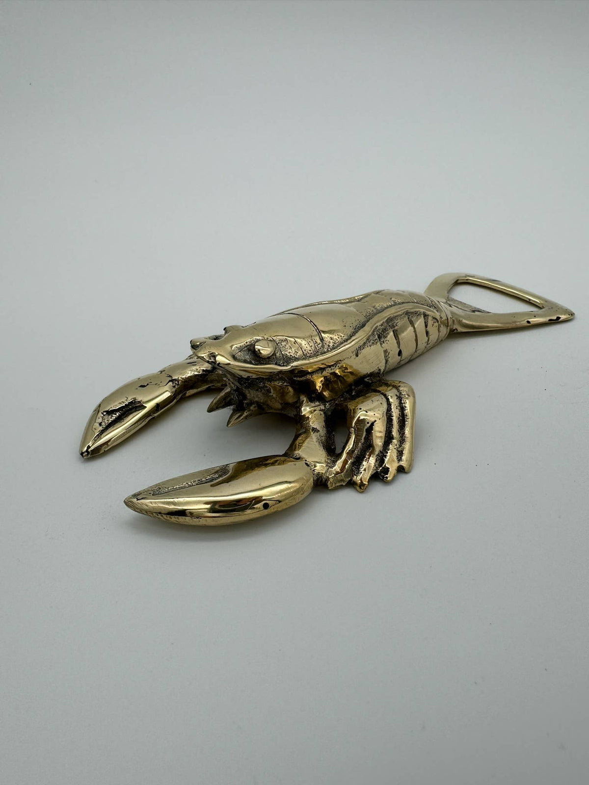A metallic lobster-shaped bottle opener with a polished gold finish, photographed on a neutral light gray background.