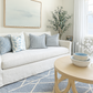 A bright, modern living room featuring a white slipcovered sofa with blue and white patterned pillows, a light wood coffee table, a blue and white geometric rug, and a large framed abstract print on the wall.