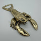 A metallic lobster-shaped bottle opener with a polished gold finish, photographed on a plain light gray background.