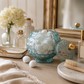 Decorative glass container with cotton wool balls on a wooden surface, surrounded by flowers and a mirror.