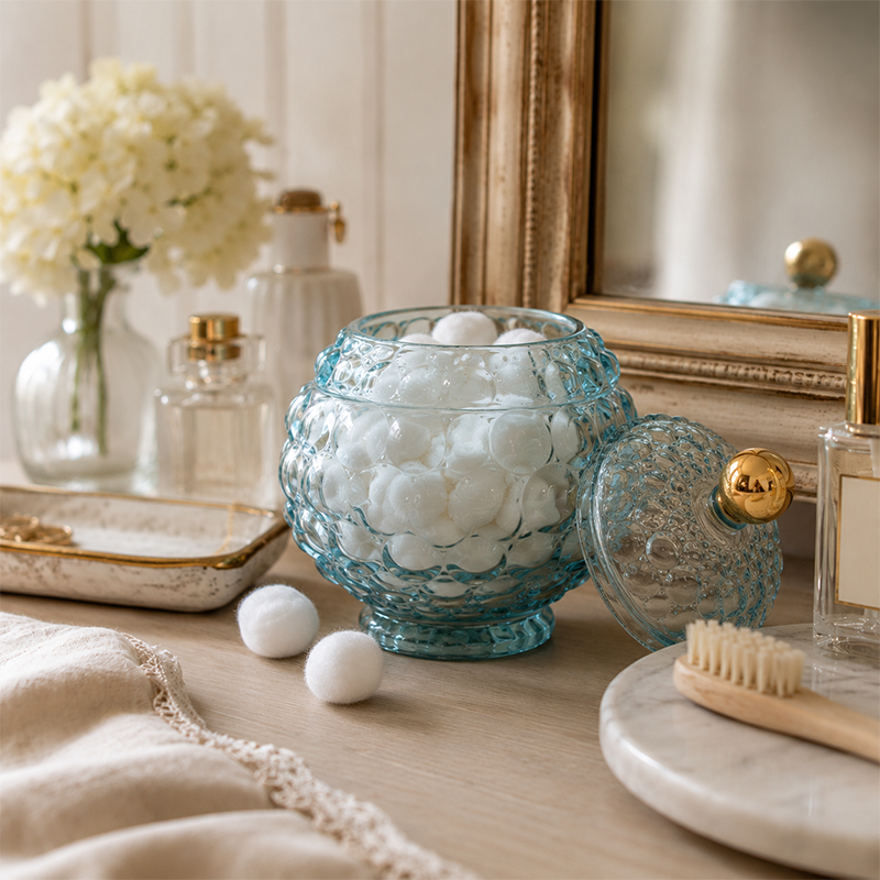 Decorative glass container with cotton wool balls on a wooden surface, surrounded by flowers and a mirror.