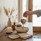 A hand holds a heart-shaped wooden keyring with several keys in a warm, styled entryway. The scene includes a rustic wooden table with a ceramic bowl, folded napkins, a candle, and dried grasses in a vase.