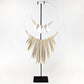 A statement necklace displayed on a black stand against a white background. The necklace features a large white circular ring with a fringe of beige wooden paddle-like elements, accented by small beige beads and three white teardrop shell pendants.