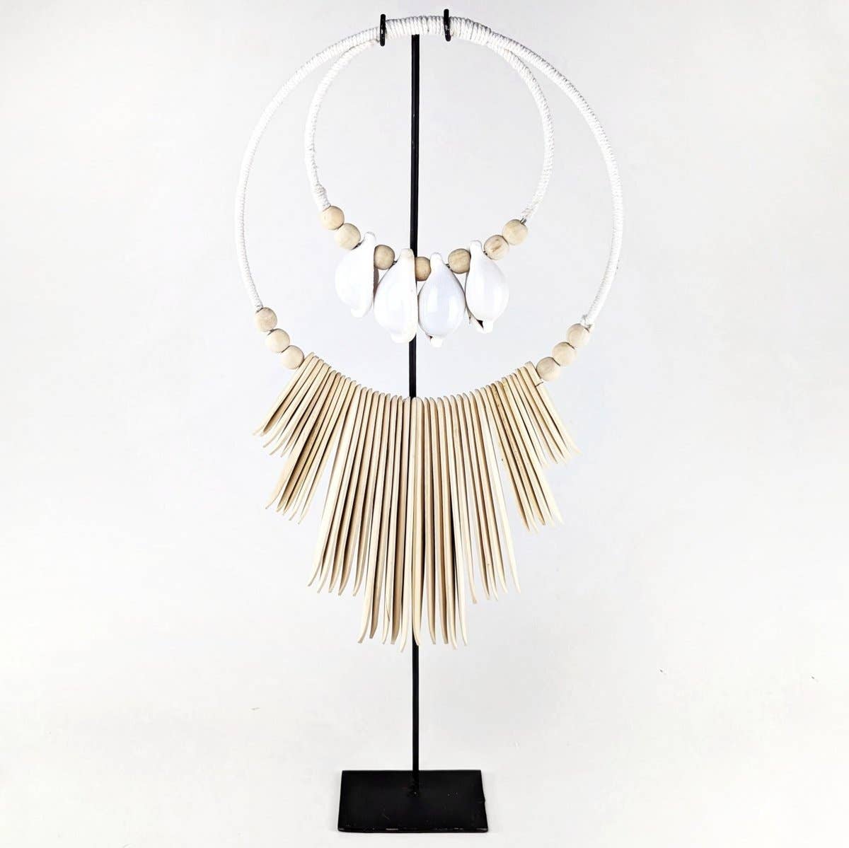 A statement necklace displayed on a black stand against a white background. The necklace features a large white circular ring with a fringe of beige wooden paddle-like elements, accented by small beige beads and three white teardrop shell pendants.