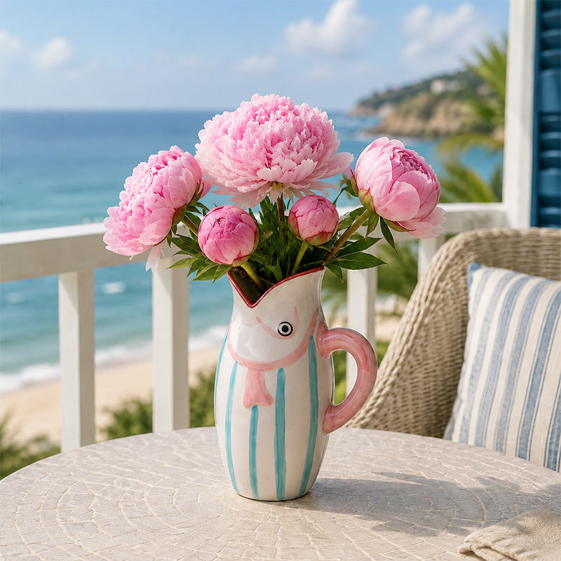 Pink peonies in a blue striped fish vase on a table with ocean view