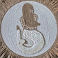 A circular woven wall hanging or rug featuring a stylized mermaid design made from natural jute rope, cowrie shells, and small seashells, set against a circular white woven center and a natural straw border.