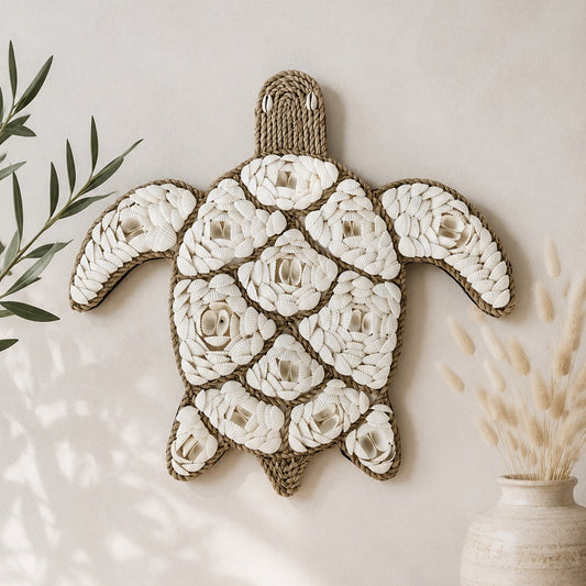 A handmade sea turtle wall hanging made from white rope flowers and natural jute trim, displayed on a light beige wall above a wooden surface with a shell, books, and a vase.