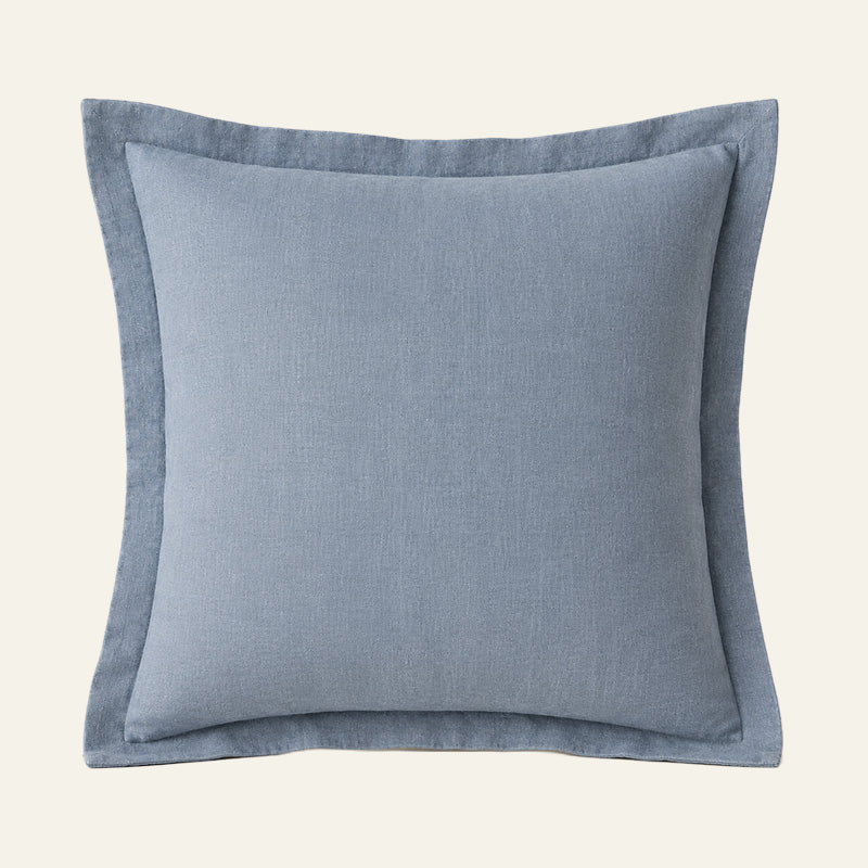A square throw pillow in a dusty light blue linen fabric with a matching piped border.