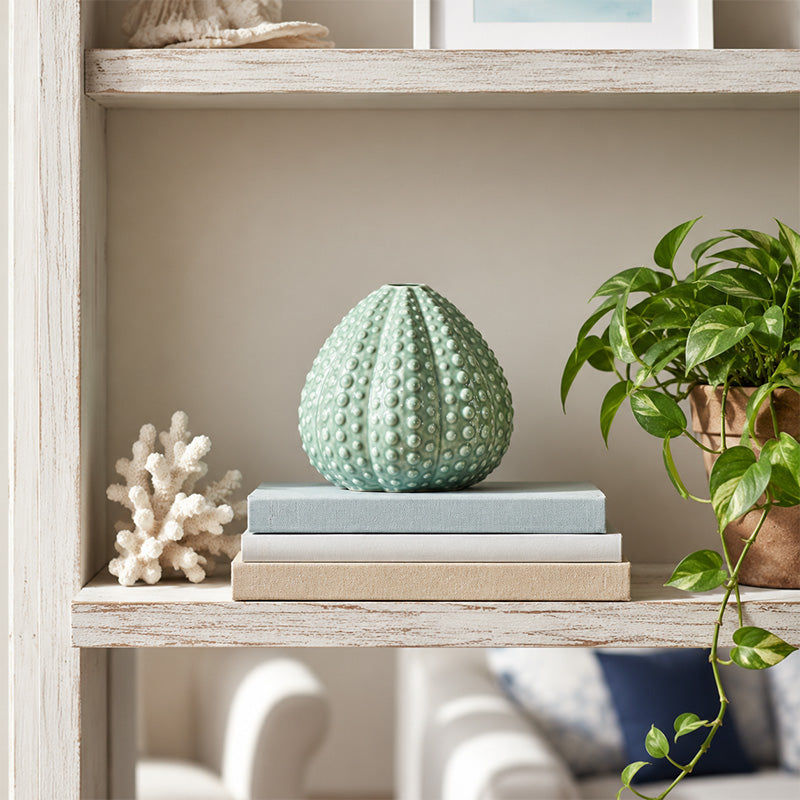 Decorative green ceramic sea urchin sculpture on books with a plant and coral in the background.