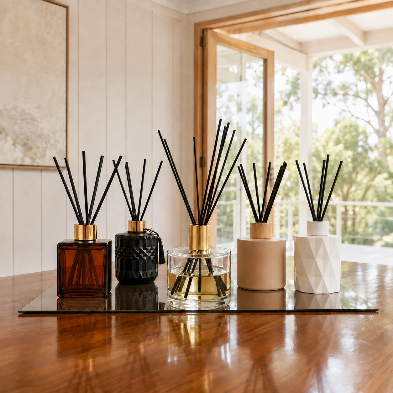 Five reed diffusers with black reeds arranged on a glass tray over a polished wooden table, with a bright room and window in the background.