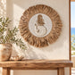 Round bohemian wall decor featuring a carved mermaid design in the center, surrounded by a natural straw raffia fringe, displayed on a cream wall above a rustic wooden console table with a potted olive branch.