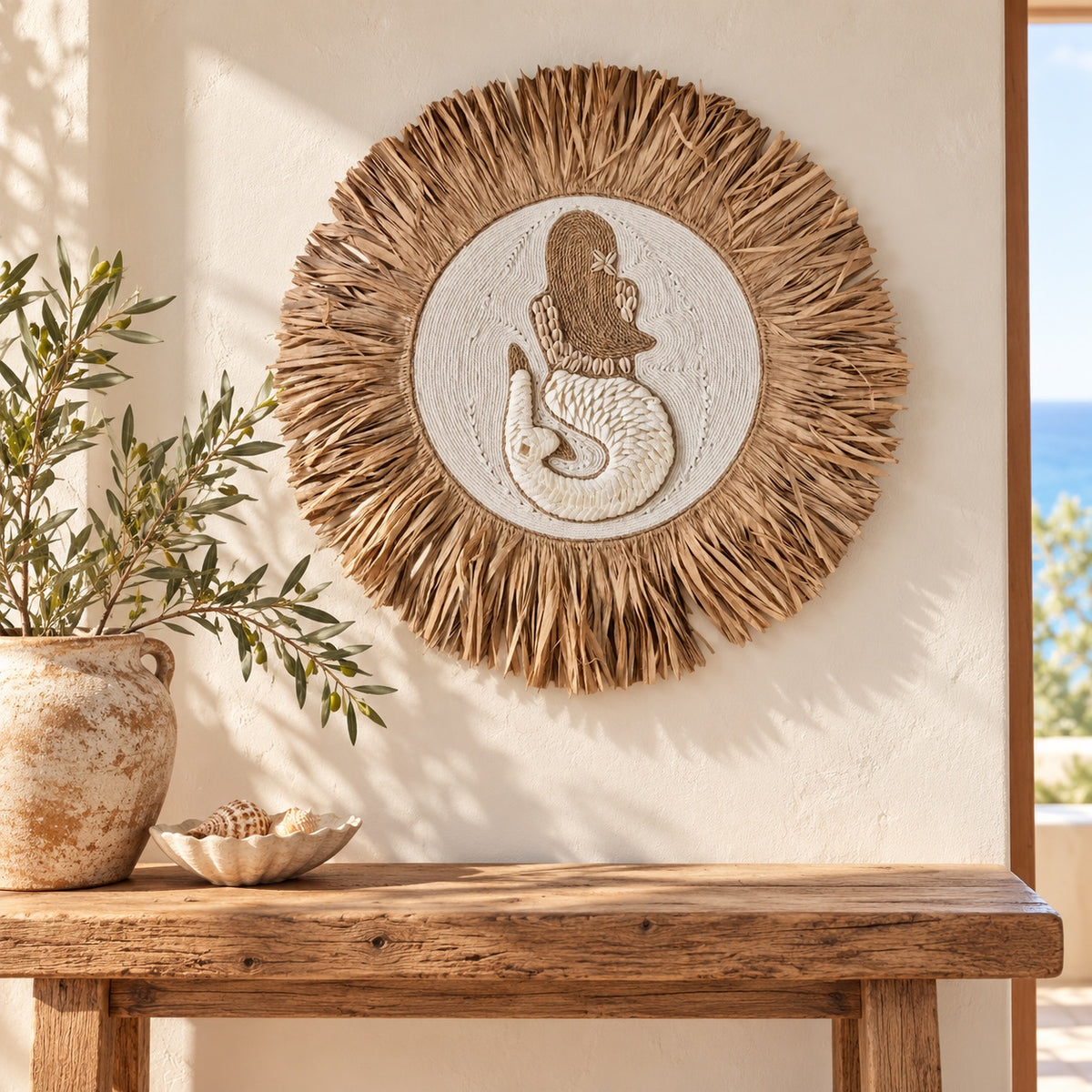 Round bohemian wall decor featuring a carved mermaid design in the center, surrounded by a natural straw raffia fringe, displayed on a cream wall above a rustic wooden console table with a potted olive branch.