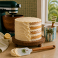 Layered cake on a wooden stand with a pot with a palm tree on it and 2 spatuals - one striped, one with a palm tree on.