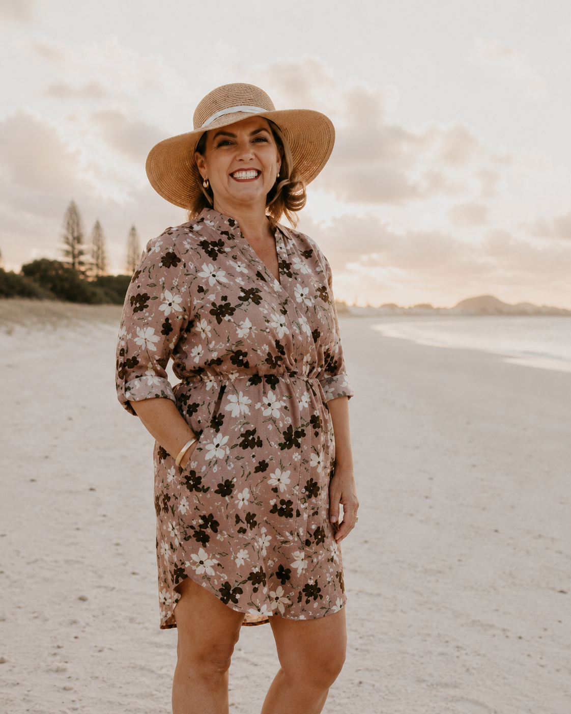 Woman in a floral dress and sun hat standing on a beach with a scenic background.