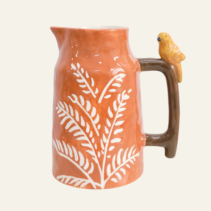 Terracotta-orange stoneware ceramic jug with white fern pattern, glossy brown handle and small yellow bird perched on top.

