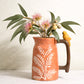 Terracotta-orange stoneware ceramic jug with white fern pattern, glossy brown handle and small yellow bird perched on top containing Australian Native flowers
