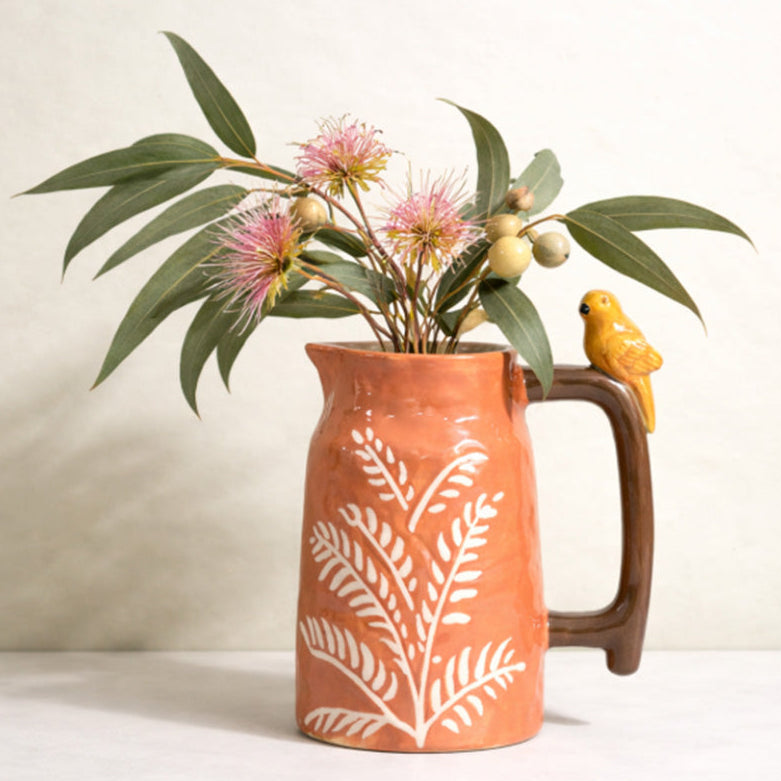 Terracotta-orange stoneware ceramic jug with white fern pattern, glossy brown handle and small yellow bird perched on top containing Australian Native flowers
