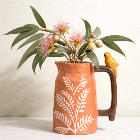 Terracotta-orange stoneware ceramic jug with white fern pattern, glossy brown handle and small yellow bird perched on top containing Australian Native flowers
