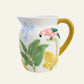Hand-painted white ceramic jug with a pink parrot, blue flowers, tropical green leaves and a mustard-yellow handle.