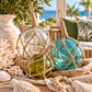 Large and medium decorative glass rope balls in Clear, Azure Aqua and Olive, wrapped in natural jute rope and styled in a rustic wooden bowl with seashells. Nautical coastal home décor from Bango Jango Australia.