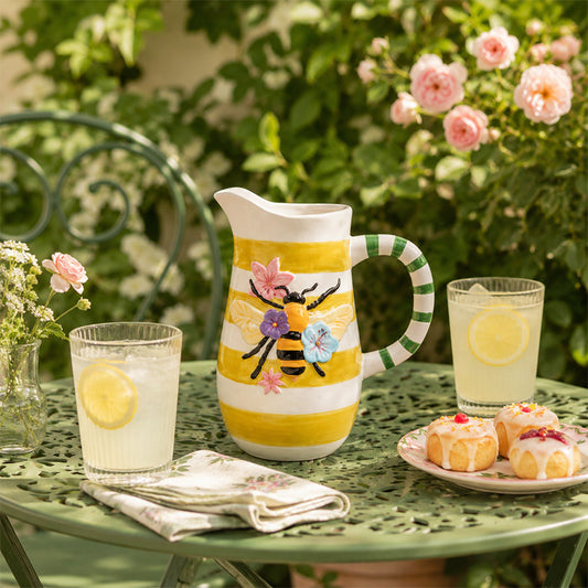Decorative pitcher with floral and bee design on a garden table with lemonade and pastries.