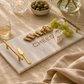 Marble cheese board with gold accents, cheese, crackers, and grapes on a beige surface.