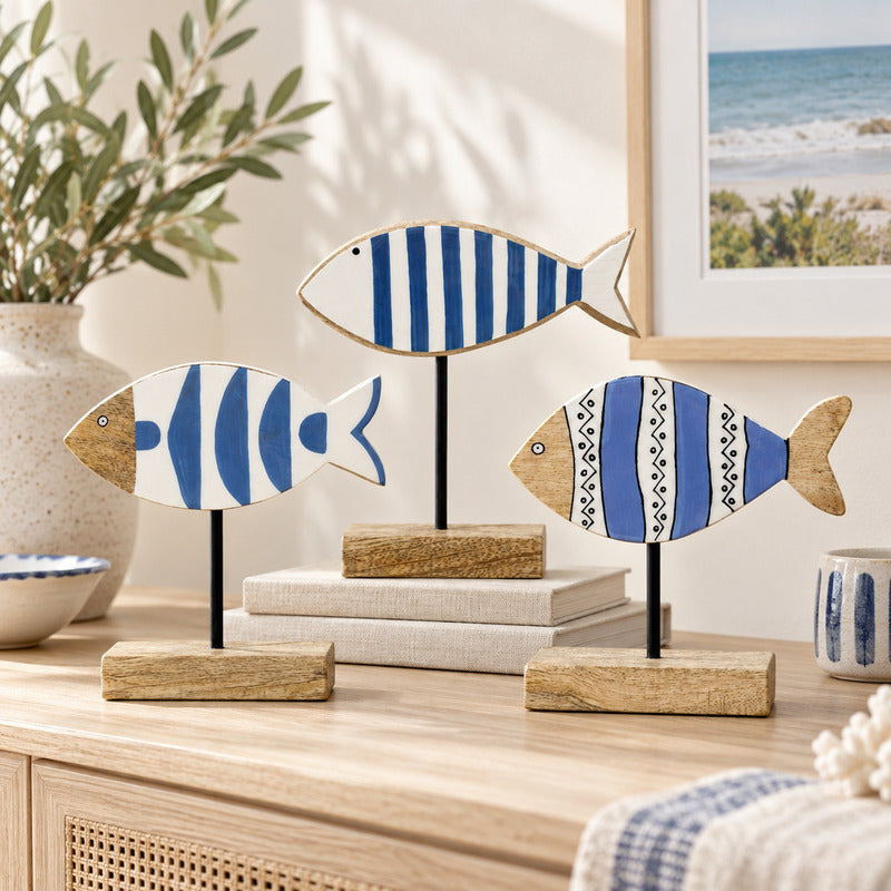 Three decorative fish-shaped wall art pieces with blue and white striped and patterned designs, mounted on black stands with wooden bases, displayed on a light wood console in a styled interior setting.