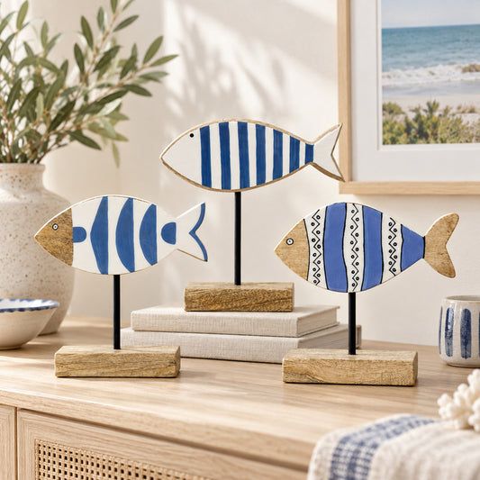 Three decorative fish-shaped wall art pieces with blue and white striped and patterned designs, mounted on black stands with wooden bases, displayed on a light wood console in a styled interior setting.