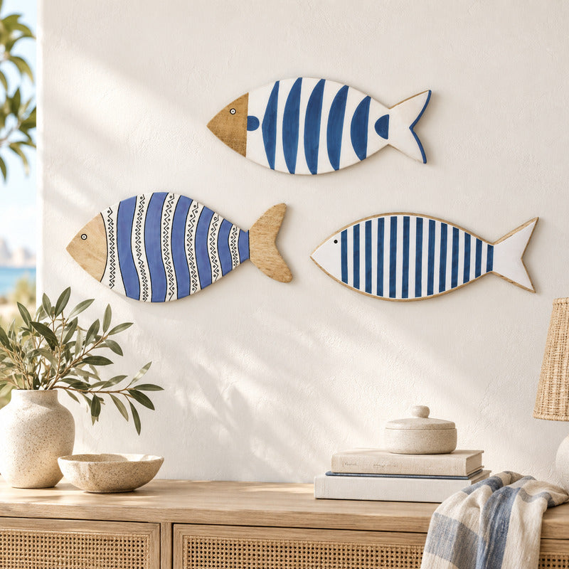 Three decorative fish wall art pieces with blue and white striped patterns and natural wood edges, displayed on a cream wall above a wooden console table with books, a vase, and a throw blanket.