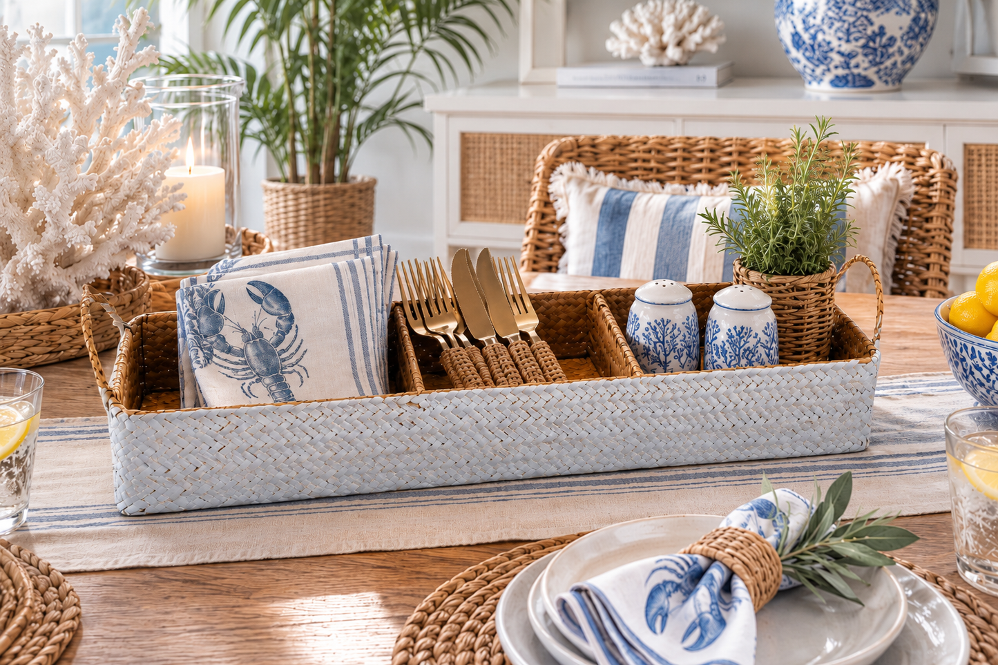 A styled table setting featuring a large woven tray holding blue-and-white lobster-print napkins, gold-toned cutlery, small blue-and-white ceramic shakers, and a small potted green plant, with matching home decor in the background.