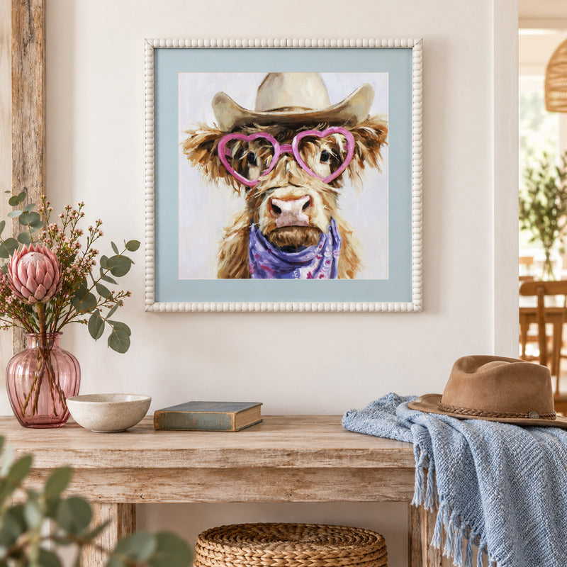 A framed art print featuring a cute Highland cow wearing pink heart-shaped sunglasses and a purple bandana, hanging on a white wall above a rustic wooden table styled with a pink vase, a blue throw blanket, and a brown hat.