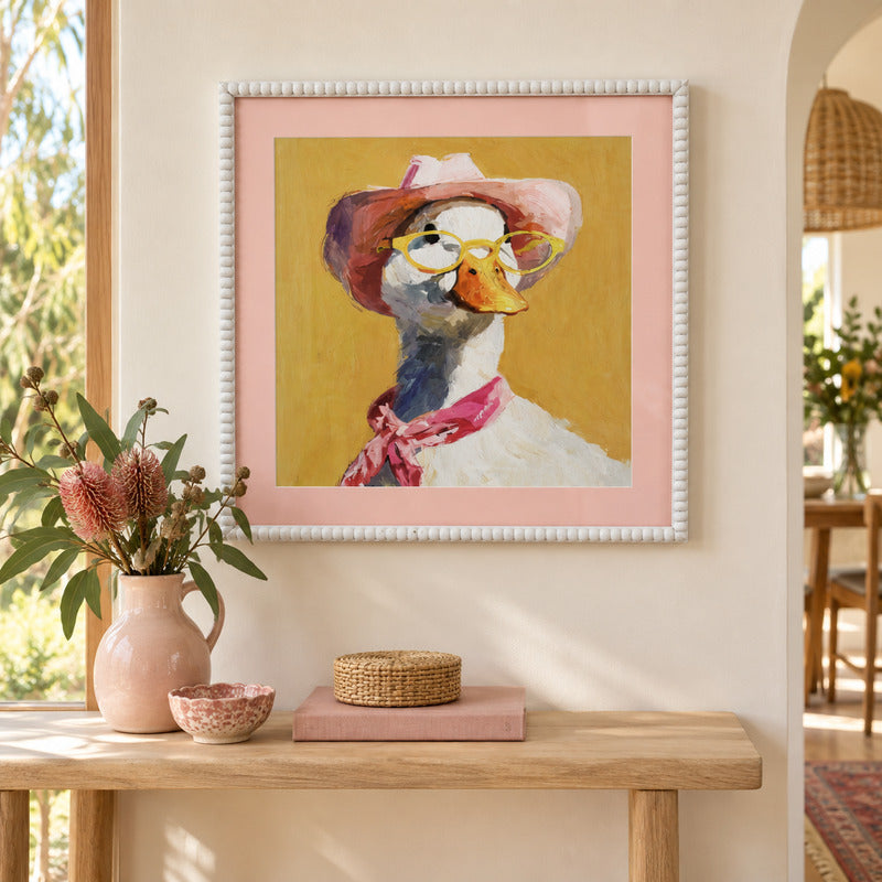 A framed art print featuring a whimsical painting of a duck wearing a pink hat, yellow glasses, and a pink scarf, displayed on a beige wall above a wooden console table with decorative items.