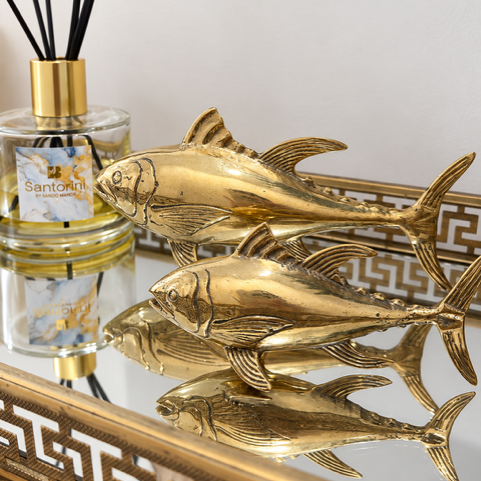 A decorative gold fish figurine displayed on a mirrored surface next to a Santoral reed diffuser bottle, with a gold-trimmed tray in the background.