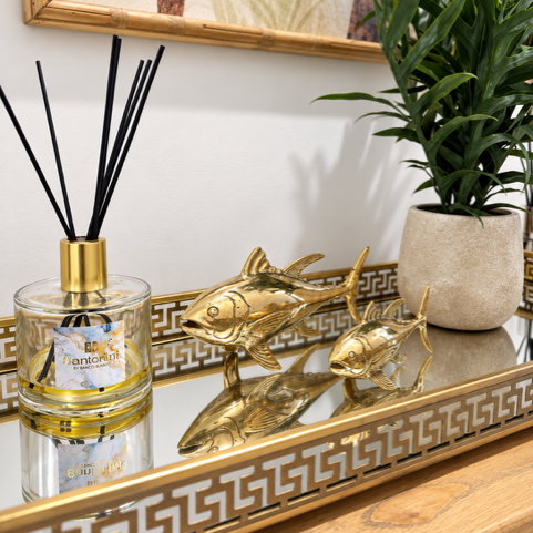 A gold decorative tray with a Greek key pattern sits on a wooden console table, styled with a reed diffuser, two gold fish sculptures, and a potted green plant. A framed botanical print hangs on the white wall behind.