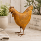 Rust-colored chicken sculpture on a wooden surface with plants in the background