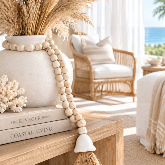 A beaded garland made of small round natural wood beads, with a white shell-shaped connector holding a brown raffia tassel, photographed on a plain white background.