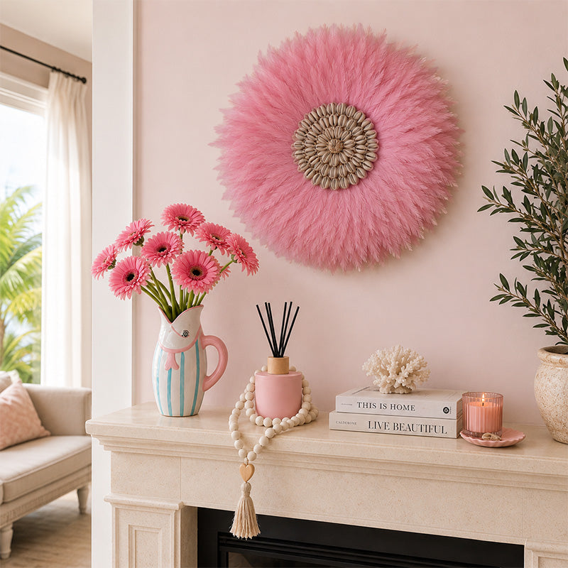A styled interior scene with a pink feather circular wall decor piece hanging on a blush pink wall above a cream fireplace mantel. The mantel holds pink gerbera daisies in a pastel pitcher, a pink reed diffuser, books, a coral ornament, a tassel garland, and a lit pink candle.