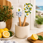 Two spatulas with lemon design in a white container on a kitchen counter with lemons and plants.