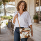 A smiling woman wearing a white button-down shirt and blue jeans carries a brown and white argyle patterned crossbody bag with tan leather trim on a sunny porch.