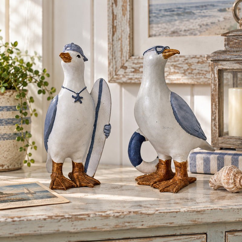 Two ceramic seagull figurines styled on a distressed white wooden table. One gull holds a blue-and-white surfboard, and both have blue wings, brown feet, and orange beaks.