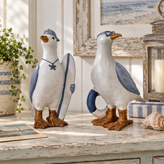 Two ceramic seagull figurines styled on a distressed white wooden table. One gull holds a blue-and-white surfboard, and both have blue wings, brown feet, and orange beaks.
